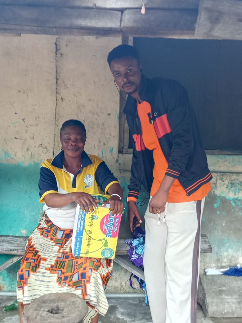 An elderly woman seated outdoors holds a carton of relief food while a volunteer stands beside her.