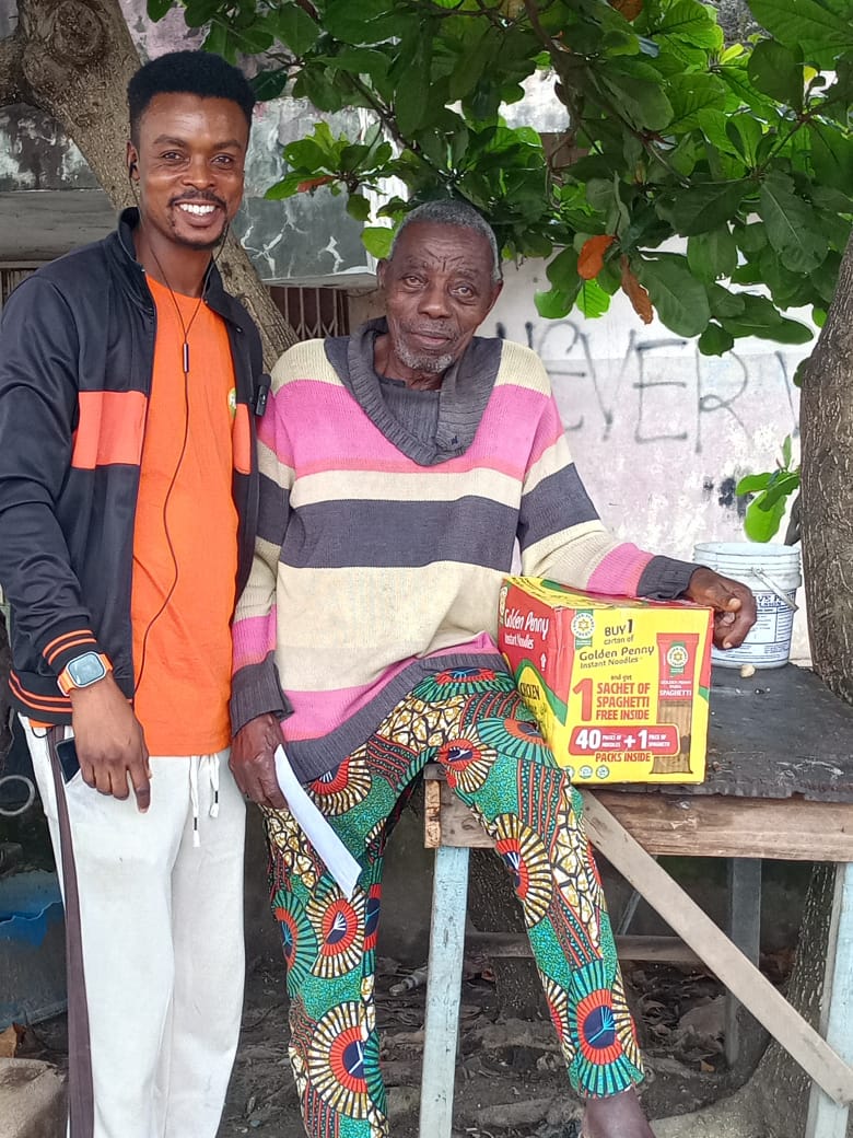 A volunteer stands beside an elderly man seated under a tree, a carton of relief food on the table beside him.