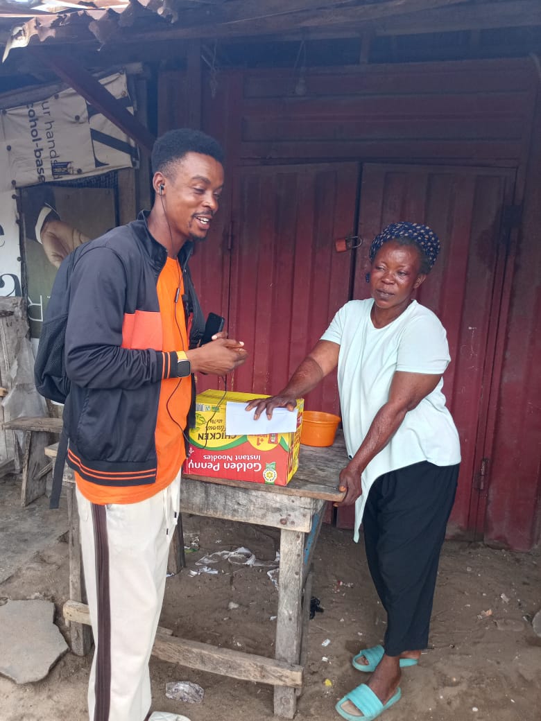 A volunteer and an elderly woman share a laugh over a carton of food supplies set on a wooden table.