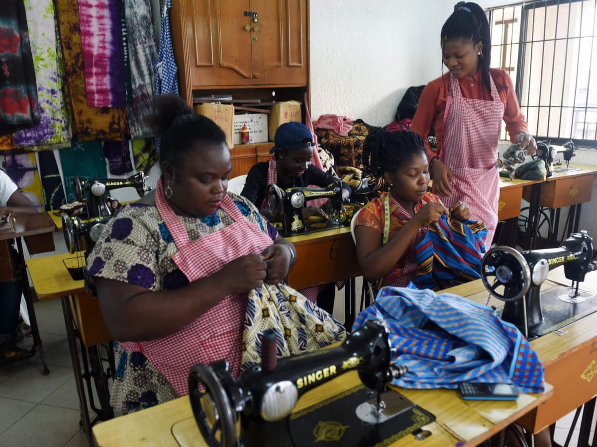 Women in gingham aprons work Ankara fabric at a row of Singer sewing machines.