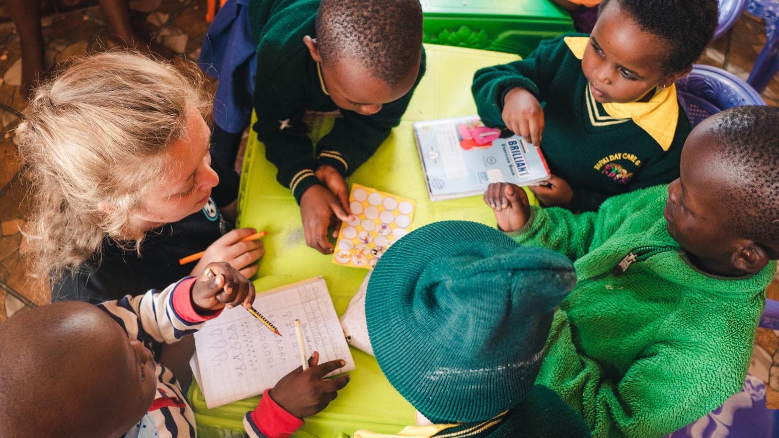Young children in green school uniforms gather round a table with a volunteer, tracing letters in exercise books.