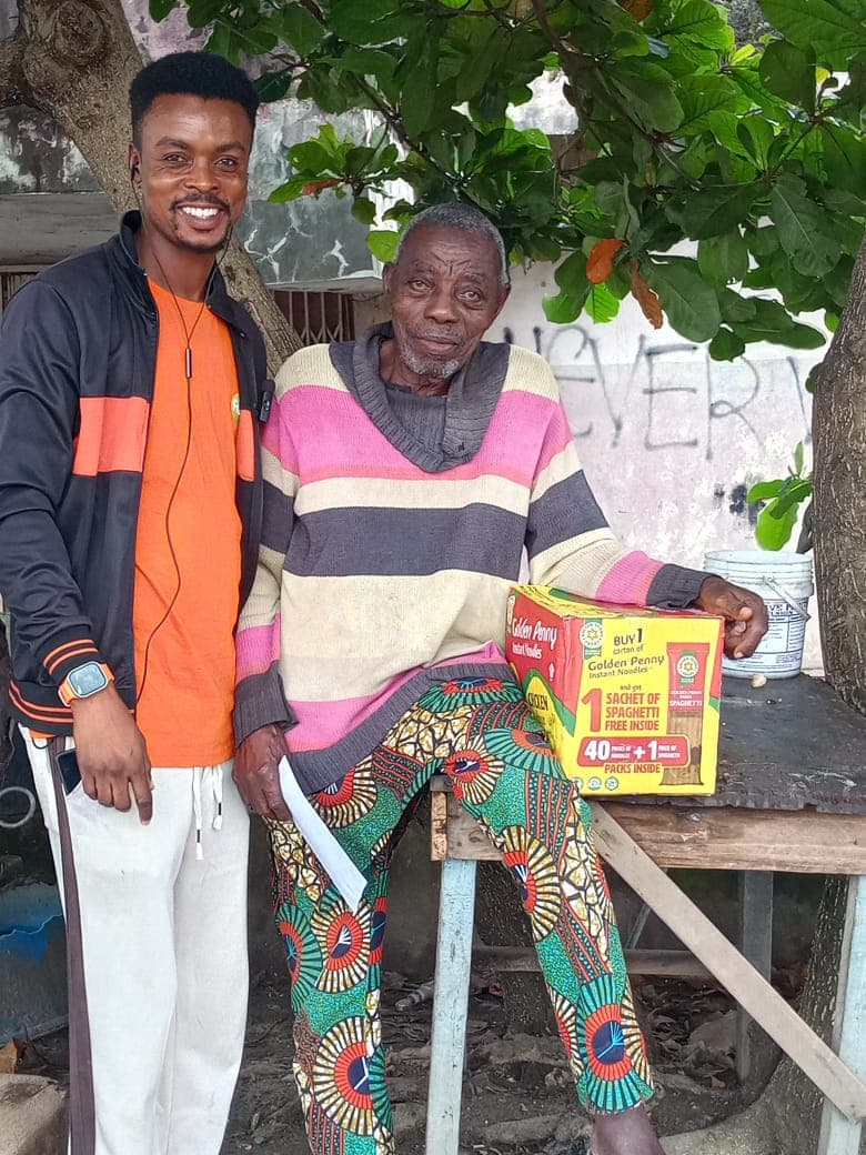 A volunteer stands beside an elderly man seated under a tree, a carton of relief food on the table beside him.